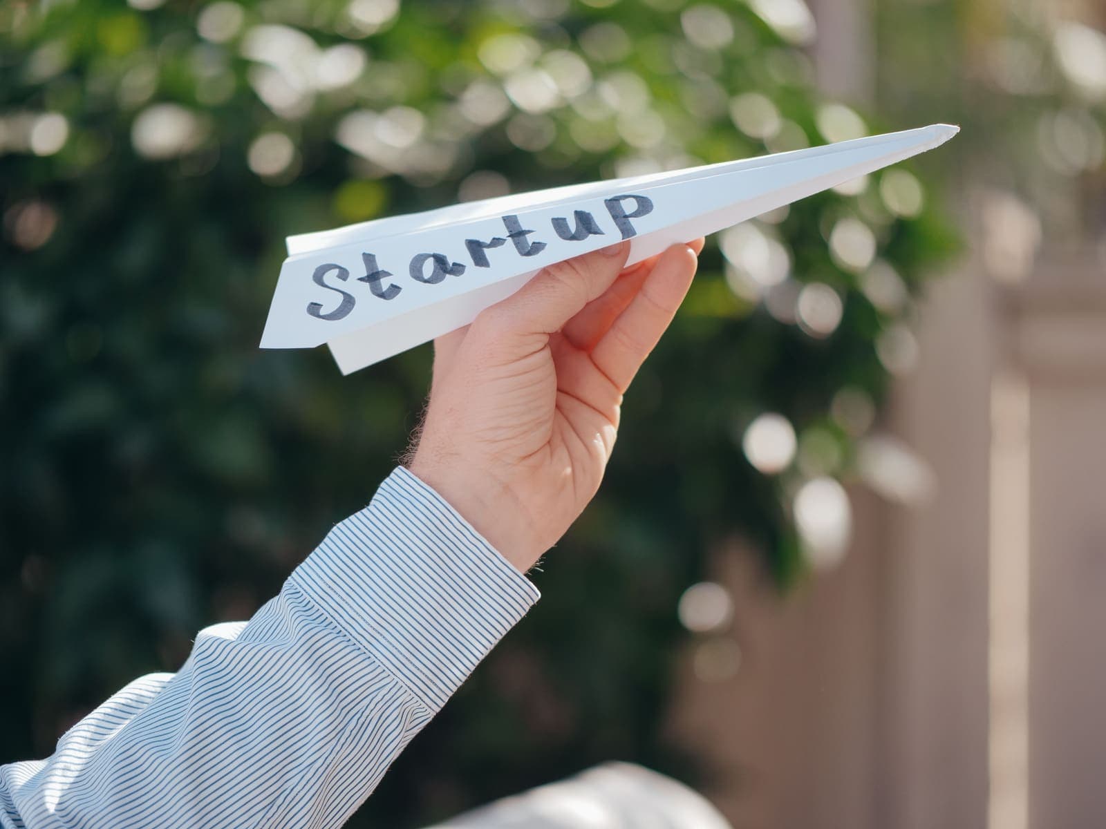 Hand launching a paper airplane labeled Startup against a green outdoor backdrop — symbolizing entrepreneurial ambition and new beginnings